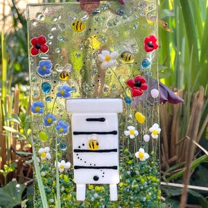 May include: A white glass beehive with black stripes and a bee on the front, surrounded by colorful flowers and bees on a green background. The beehive is on a wooden base.