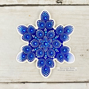 May include: A blue and white snowflake sticker with a mandala design. The sticker is on a white wooden background. Delicate Dots Andrea Moebes ©