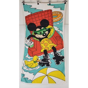 May include: A white beach towel featuring a graphic of Mickey Mouse wearing green goggles and yellow patterned swim trunks while lounging in a red inflatable chair. The scene includes a yellow rubber duck, a beach ball, and a drink.