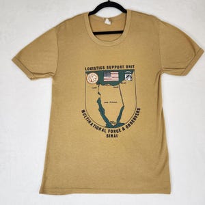 May include: A tan short-sleeve t-shirt featuring a graphic print of the Sinai Peninsula map. The text reads Logistics Support Unit, Multinational Force & Observers, and Sinai. It includes a small American flag and two circular unit emblems.