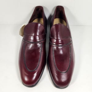 May include: A pair of dark burgundy leather loafers featuring a polished, glossy finish, a classic penny slot strap across the vamp, and a rounded toe design. The shoes have a smooth, structured silhouette with visible dark stitching along the edges.