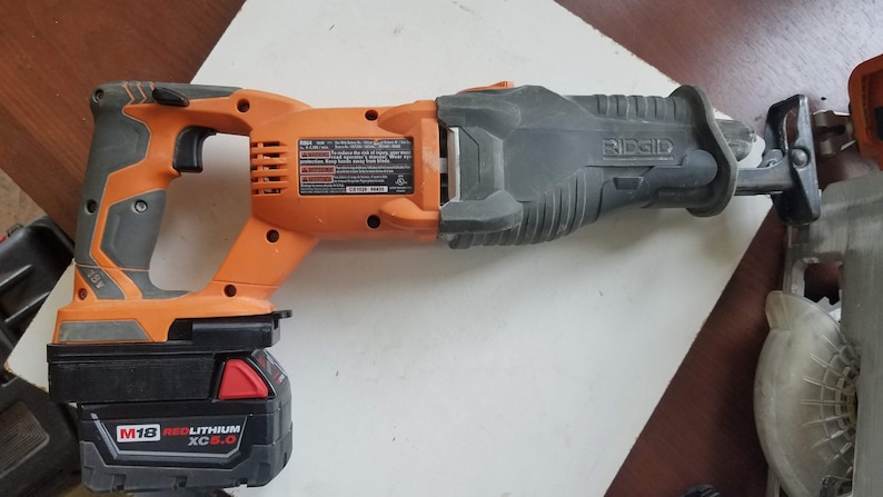 May include: An orange and black cordless reciprocating saw with a black battery pack labeled "M18 RED LITHIUM XC 6.0" and the brand name "RIDGID" on the saw.
