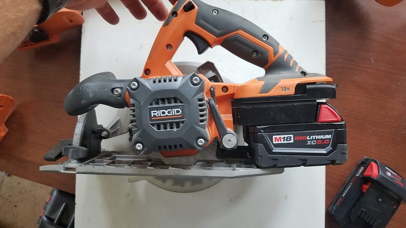 May include: An orange and black cordless circular saw with a 7-1/4 inch blade. The saw has a black battery pack labeled "M18 REDLITHIUM XC5.0" and a black and orange handle. The saw is made by RIDGID.