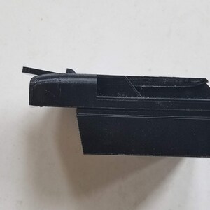 May include: A black plastic 3D printed object with a rectangular shape and a small, angled flap on the top.