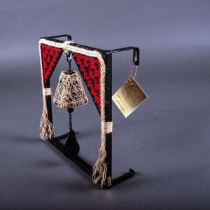 May include: A black metal bell with a woven rope design and red accents hangs from a black metal frame. The bell is attached to a chain and has a woven rope tassel. A small tag is attached to the frame.