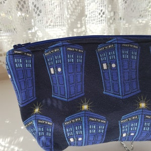 Puede incluir: Una bolsa azul con cremallera con un patrón repetido de cajas de policía azules con el texto "Police Box" en ellas. La bolsa está hecha de tela y tiene una cremallera.