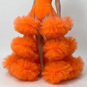 Puede incluir: Un body naranja brillante sin mangas con cuello alto y una falda superpuesta desmontable. La falda está confeccionada con múltiples niveles de tela de tul voluminosa y con volantes, creando una apariencia texturizada y en capas.