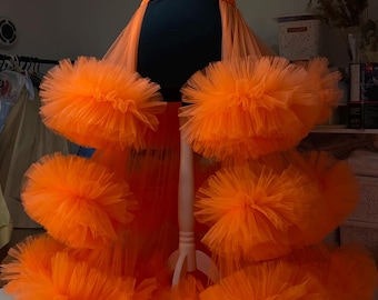 May include: A bright orange tulle skirt with multiple layers of ruffles. The skirt is on a mannequin and has a black waistband.