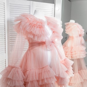 May include: A pink tulle robe with a ruffled collar and sleeves. The robe has a bow at the waist and is layered with ruffles.