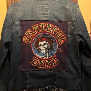 May include: A denim jacket with a large patch on the back featuring a skull surrounded by roses and the words "Grateful Dead" in a circular design. The jacket is a medium wash blue.