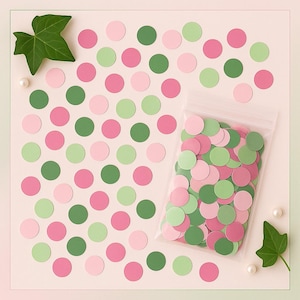 May include: A collection of pink, green, and light pink confetti circles scattered on a light pink surface. A clear plastic bag filled with similar confetti is on the right. Green leaves and pearls are placed around the confetti.