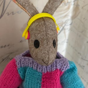 May include: A handcrafted bunny doll with a grey body, long ears, and black button eyes. The bunny is wearing a colourful knitted sweater in pink, blue, and purple, and yellow headphones. A whimsical toy.