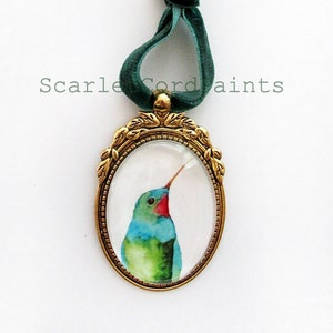 May include: A gold-toned oval pendant with a green velvet ribbon. The pendant features a watercolor painting of a hummingbird with blue, green, and red colors.