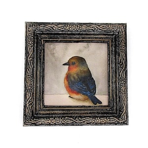 May include: A framed watercolor painting of a small bird with orange, blue, and brown feathers. The bird is perched on a branch and looking to the left. The frame is black with gold accents.