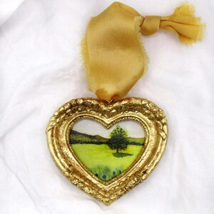 May include: A gold heart-shaped frame with a small painting of a green field and a lone tree inside. The frame is attached to a gold ribbon.