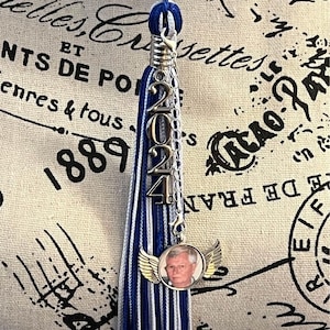May include: A blue, white, and silver graduation tassel with a 2024 charm and a silver charm with a photo of a man wearing a white shirt and a silver chain. The tassel is hanging from a graduation cap.