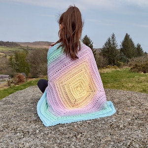 May include: A person wearing a rainbow crochet blanket with a gradient of colors from blue to purple. The blanket is crocheted with a delicate stitch pattern and has a square design in the center.