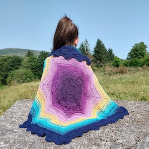 Puede incluir: Un chal de ganchillo colorido con un degradado de azul, amarillo, rosa y morado. El chal está hecho con un patrón de estrella y tiene un borde festoneado.
