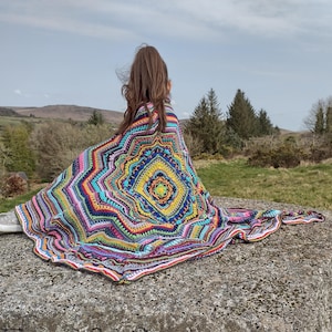 Könnte beinhalten: Eine farbenfrohe, handgefertigte Häkeldecke mit geometrischem Muster. Die Decke zeigt eine lebendige Farbpalette, darunter Blau, Gelb, Rosa und Grün. Die Decke ist über einer Person drapiert, die auf einer Steinoberfläche sitzt, mit einer Naturlandschaft im Hintergrund.