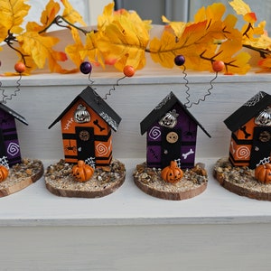 May include: Four miniature Halloween houses with black roofs, orange and purple painted walls, and black doors. Each house has a small pumpkin and other Halloween decorations on the front.