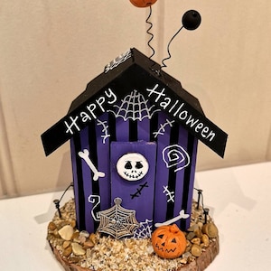 May include: A purple and black Halloween house with a Jack Skellington face on the door. The house has a black roof with the words "Happy Halloween" written on it. There are two orange pumpkins on the roof and a spider web on the side of the house.