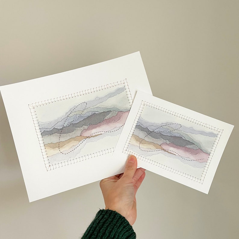 May include: Two framed watercolor paintings of abstract landscapes in shades of gray, pink, and beige. The paintings are stitched with black thread around the edges.
