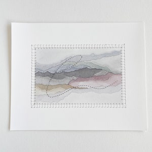 May include: A watercolor painting of a landscape with a stitched border. The painting features a series of layered hills in shades of gray, brown, and pink. A black dotted line forms a curved shape in the center of the painting.