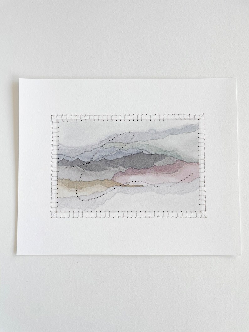 May include: A watercolor painting of a landscape with a stitched border. The painting features a series of layered hills in shades of gray, brown, and pink. A black dotted line forms a curved shape in the center of the painting.