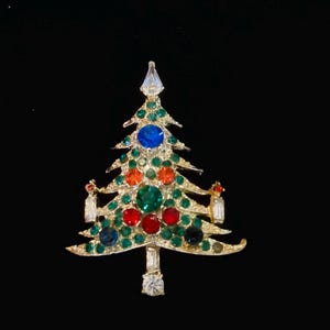 May include: A gold-tone metal Christmas tree brooch featuring a variety of colorful rhinestones. The tree is decorated with small green stones, larger blue, red, and orange circular stones, and clear baguette-cut stones at the base and sides.