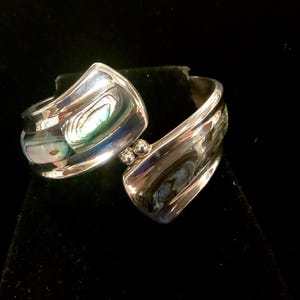 May include: A silver-toned cuff bracelet with an open design. The bracelet features iridescent abalone shell inlays and two small silver spheres at the clasp. The polished metal reflects light, creating a striking contrast.