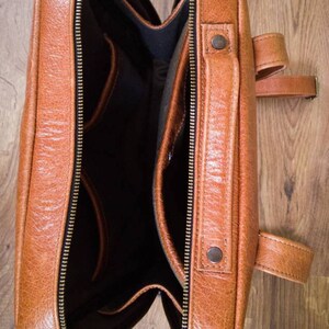 K&ouml;nnte beinhalten: Offener brauner Lederrucksack mit mehreren Taschen und einem Rei&szlig;verschluss. Der Rucksack hat eine Klappe mit einem Schnallenverschluss.