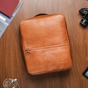 K&ouml;nnte beinhalten: Eine braune Ledertasche f&uuml;r Laptops mit Rei&szlig;verschluss. Die Tasche liegt auf einer Holzoberfl&auml;che.