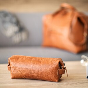 Könnte beinhalten: Eine braune Reise-Toilette-Tasche aus Leder mit Reißverschluss. Die Tasche liegt auf einem Holztisch.