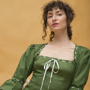 May include: A green, short-sleeved blouse with white ribbon ties and white stitching. The blouse has a square neckline and a fitted bodice. The sleeves are puffed and have a ruffle at the cuff.