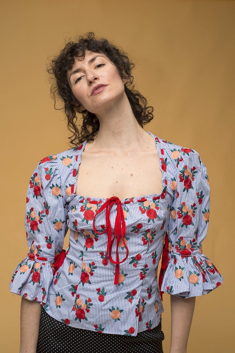 May include: A blue and white striped blouse with red embroidered flowers and a red ribbon tie at the neckline. The blouse has puffed sleeves with ruffles at the cuffs.