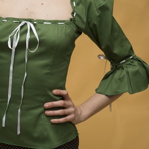 May include: A green, short-sleeved blouse with a white ribbon tie at the neckline. The blouse has a square neckline and white stitching around the edges. The sleeves are puffed and have a ruffle at the bottom. The blouse is worn over a black skirt with white polka dots.