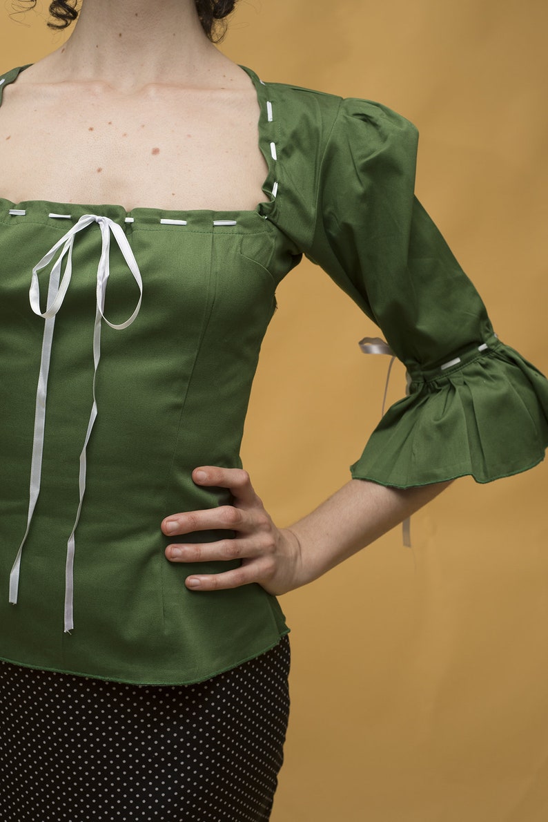 May include: A green, short-sleeved blouse with a white ribbon tie at the neckline. The blouse has a square neckline and white stitching around the edges. The sleeves are puffed and have a ruffle at the bottom. The blouse is worn over a black skirt with white polka dots.