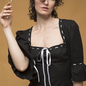 May include: A woman with curly hair is wearing a black top with white stitching and a white ribbon tied at the neckline. She is holding a dried flower in front of her eye.