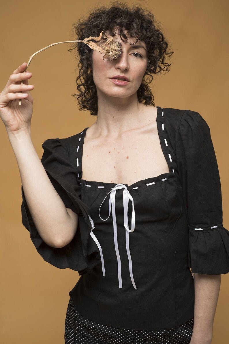 May include: A woman with curly hair is wearing a black top with white stitching and a white ribbon tied at the neckline. She is holding a dried flower in front of her eye.