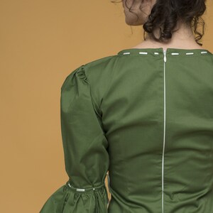 May include: A green blouse with a white zip down the back and white stitching around the neckline and sleeves. The blouse has a ruffled sleeve and a fitted bodice.