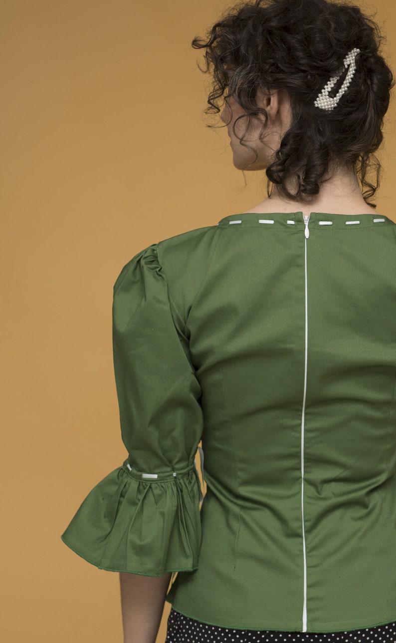 May include: A green blouse with a white zip down the back and white stitching around the neckline and sleeves. The blouse has a ruffled sleeve and a fitted bodice.