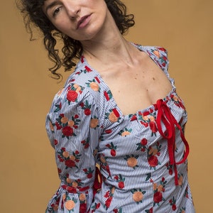 May include: A woman wearing a blue and white striped blouse with red and pink floral embroidery. The blouse has a square neckline and a red ribbon tied at the front. She is also wearing a black and white polka dot skirt.