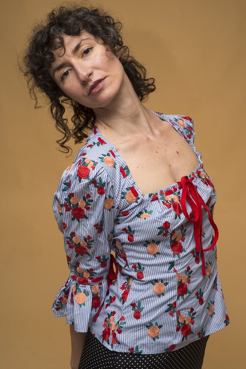 May include: A woman wearing a blue and white striped blouse with red and pink floral embroidery. The blouse has a square neckline and a red ribbon tied at the front. She is also wearing a black and white polka dot skirt.