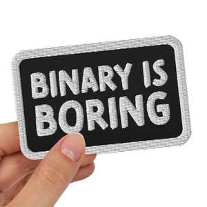Op de afbeelding: Een zwarte patch met witte geborduurde tekst die "BINARY IS BORING" luidt, vastgehouden in een hand.
