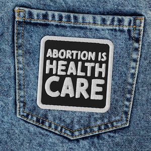 May include: A pair of blue denim jeans with a black square patch sewn onto the back pocket. The patch has white text that reads "ABORTION IS HEALTH CARE".