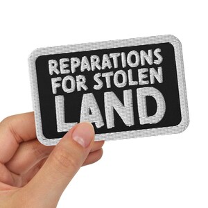 May include: A black patch with white text that reads "REPARATIONS FOR STOLEN LAND".