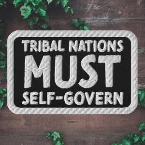 Pode incluir: Etiqueta bordada preta e branca com o texto "Tribal Nations Must Self-Govern" em um fundo branco.
