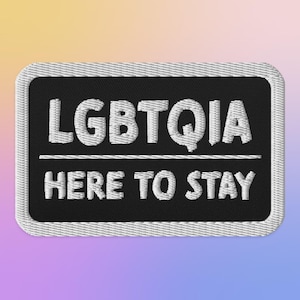 May include: A black and white embroidered patch with the text "LGBTQIA HERE TO STAY" in white lettering.