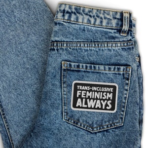 Op de afbeelding: Een paar blauwe denim jeans met een zwart-witte patch op de achterzak met de tekst "Trans-Inclusive Feminism Always".