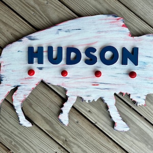 May include: A white wooden buffalo silhouette with red and blue paint accents. The name "HUDSON" is spelled out in blue letters on the buffalo. There are four red round knobs on the buffalo.